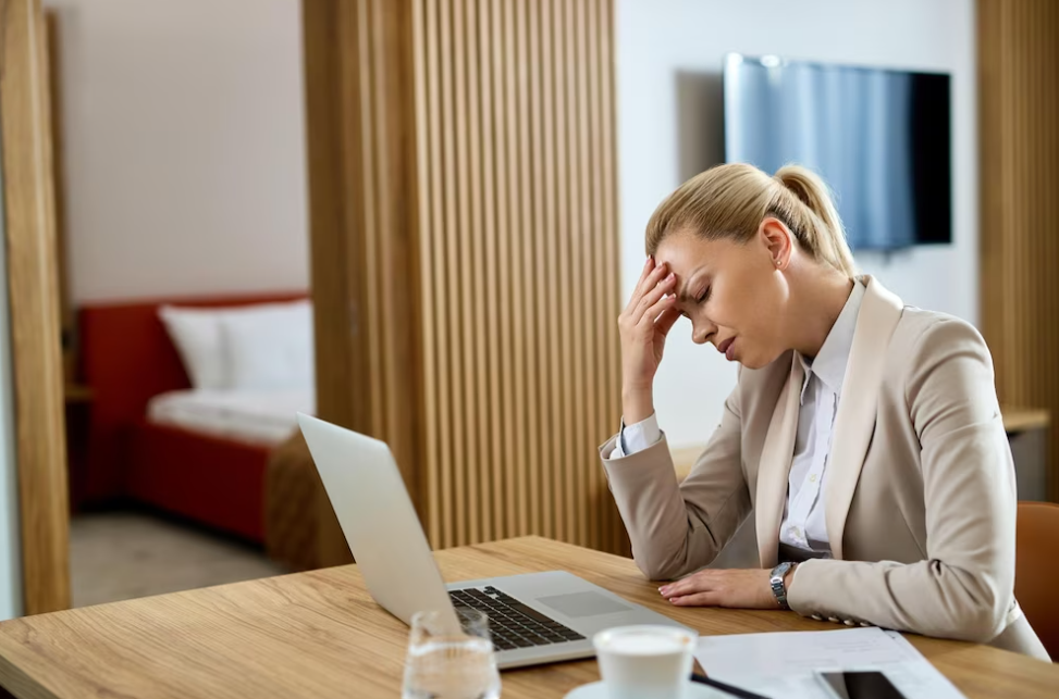 woman in a beige classic suit sitting at the desk with her hand on her forehead near the laptop at work