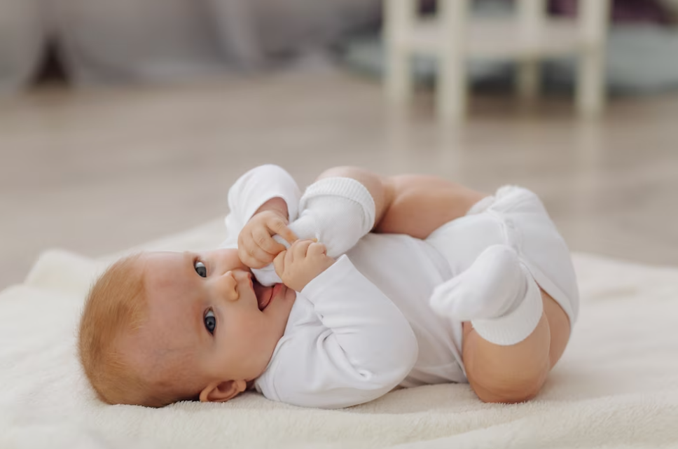 child in white clothing lying on the carpet and holds foot near opened mouth